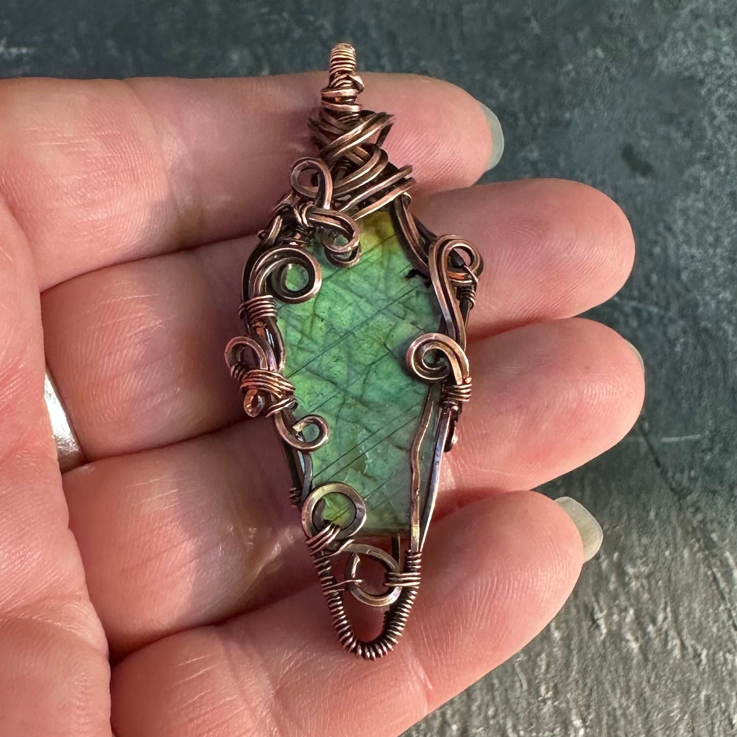 A handmade copper wire pendant with coffin shaped labradorite stone, held in a person's hand against a dark textured background.