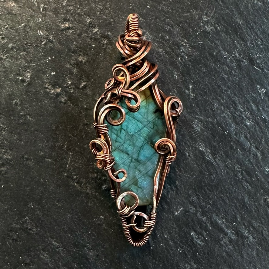 A handmade copper wire wrap pendant featuring a coffin-shaped labradorite stone, displayed on a dark textured slate background.