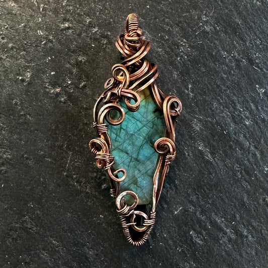 A handmade copper wire wrap pendant featuring a coffin-shaped labradorite stone, displayed on a dark textured slate background.