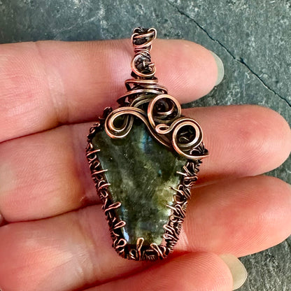 A handmade copper wire pendant wrapped around coffin shaped labradorite, held in a person's hand against a dark textured stone background.