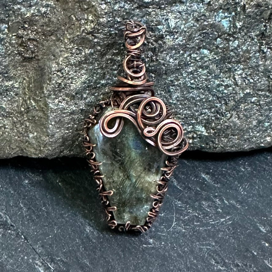 A handmade pendant featuring a coffin-shaped green labradorite stone wrapped in copper wire, set against a dark textured rock background.