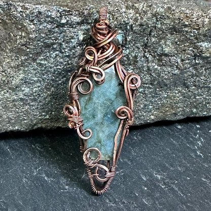 A handmade wire-wrapped pendant featuring a coffin-shaped labradorite stone, set against a dark gray slate and a larger textured gray rock background.