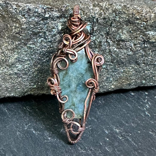 A handmade wire-wrapped pendant featuring a coffin-shaped labradorite stone, set against a dark gray slate and a larger textured gray rock background.