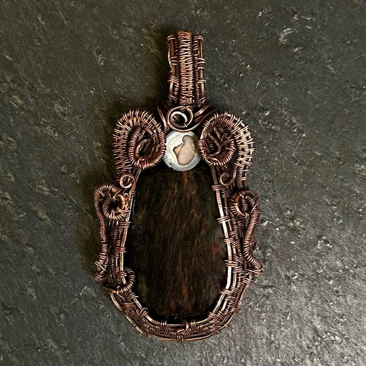 A handmade copper wire pendant with a rustic, organic coiled design and a central mahogany stone, displayed on a dark textured slate surface.