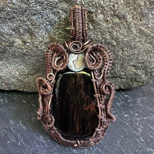A handmade wire-wrapped pendant featuring a mahogany stone centerpiece, a small white stone, and intricate copper-toned wrapping, displayed against a gray stone background.