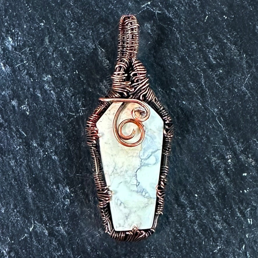 A coffin-shaped pendant featuring a white and gray Maligano japser inlay, wrapped in textured copper wire with a spiral accent on the front, set against a dark slate background.