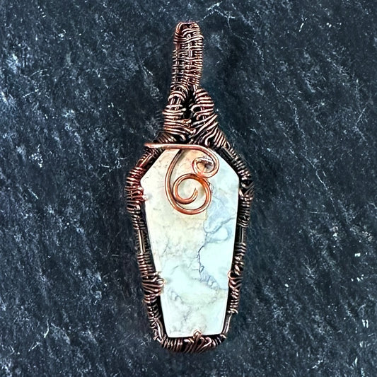A coffin-shaped pendant featuring a white and gray Maligano japser inlay, wrapped in textured copper wire with a spiral accent on the front, set against a dark slate background.