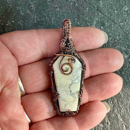 A coffin-shaped cabochon pendant with a white and gray marbled Maligano jasper center, wrapped in copper wire, held in a person's hand against a dark textured background.
