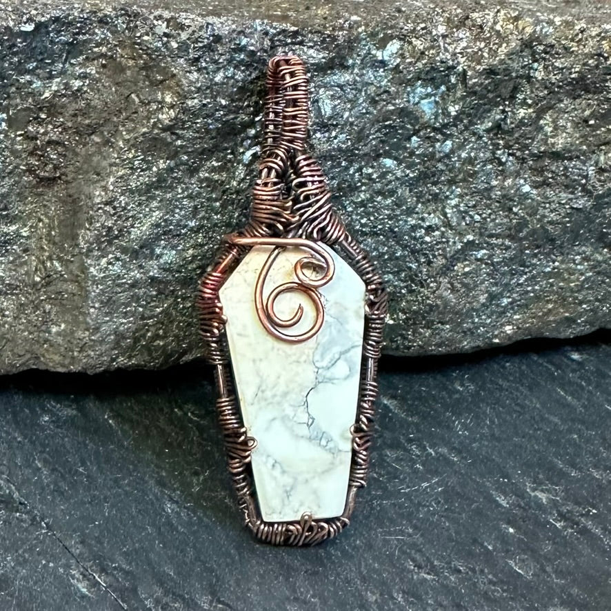 A coffin-shaped pendant featuring a white and gray Maligano stone wrapped in textured copper wire, displayed against a dark, rough-hewn rock background.
