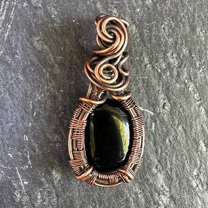 A handmade oval pendant featuring a polished black onyx stone wrapped in textured copper wire, set against a dark gray slate background.