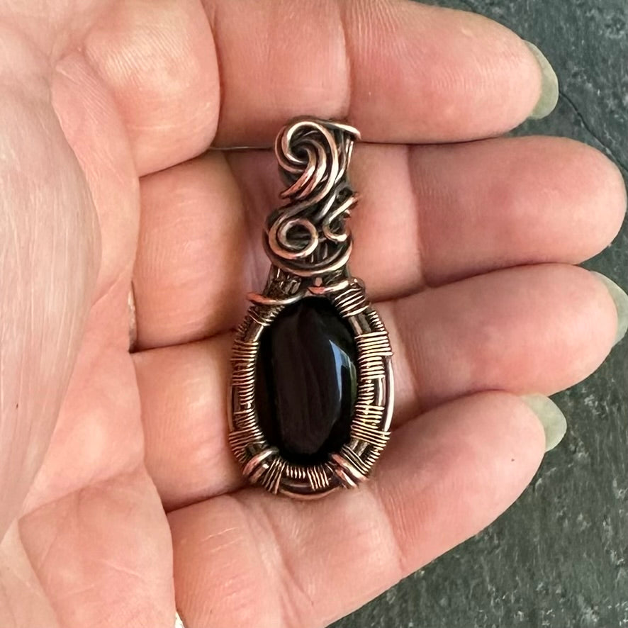 A handmade oval pendant with a black onyx stone wrapped in textured copper wire, held in the palm of a hand.