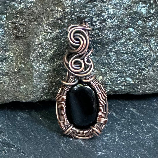 A handmade oval pendant featuring a black onyx stone wrapped in textured copper wire, set against a dark gray slate and rock background.
