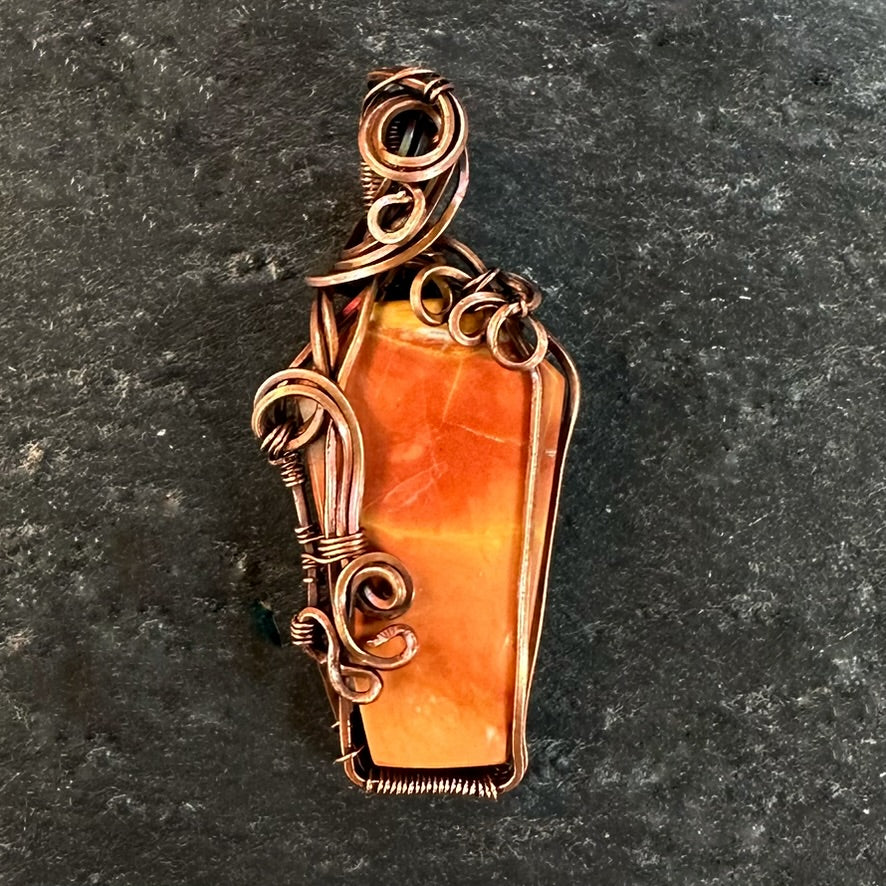 A coffin-shaped orange jasper gemstone pendant wrapped in copper wire, displayed on a dark textured slate background.