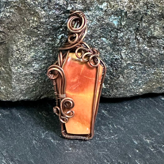 A coffin-shaped pendant featuring an  orange and peach Maligano jasper stone inlaid with copper wire wrapping, displayed against a dark textured rock background.
