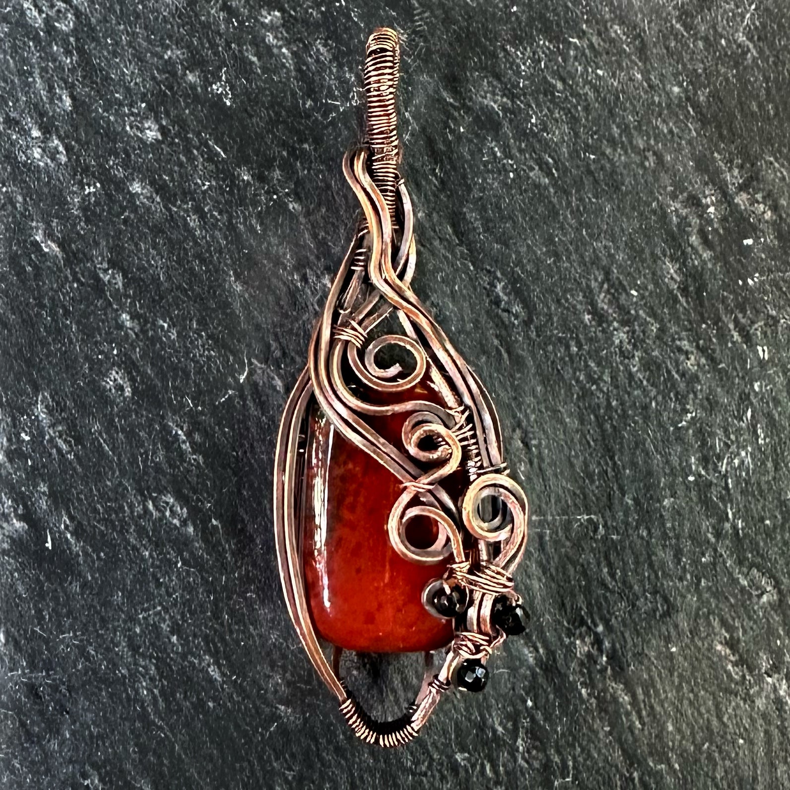 A handmade teardrop-shaped copper wire-wrapped pendant featuring a red jasper stone and small black beads, set against a dark textured slate background.