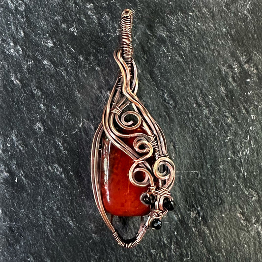 A handmade teardrop-shaped copper wire-wrapped pendant featuring a red jasper stone and small black beads, set against a dark textured slate background.