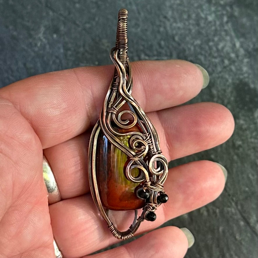 A close-up of a hand holding a teardrop-shaped pendant with a red jasper stone and intricate copper wire wrapping, set against a dark textured background.