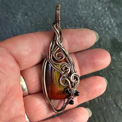 A close-up of a hand holding a teardrop-shaped pendant with a red jasper stone and intricate copper wire wrapping, set against a dark textured background.