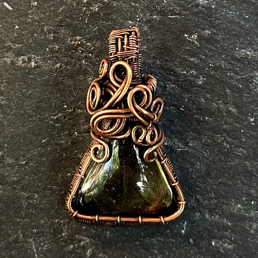 A handmade wire-wrapped pendant featuring a triangular shaman dream quartz glass stone and intricate copper wire wrapping, displayed on a dark textured slate surface.