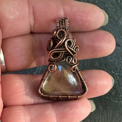 A handmade copper wire-wrapped pendant with a triangular shaman dream quartz stone, held in a person's hand.