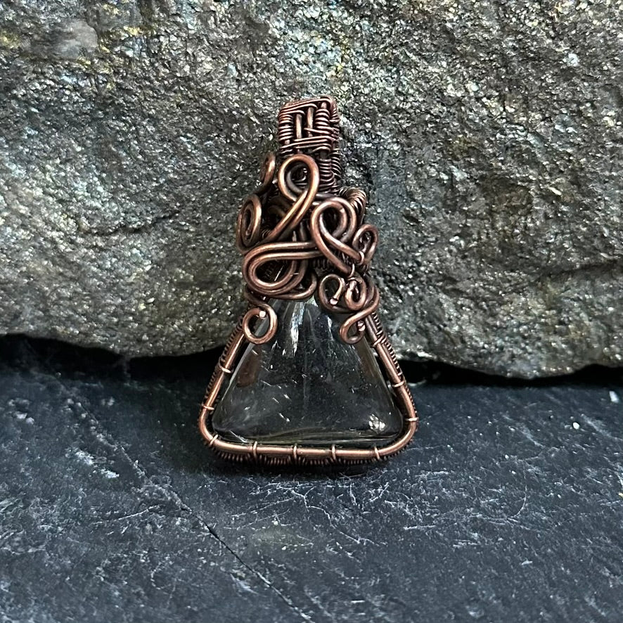 A handmade pendant featuring a shaman dream quartz wrapped in intricate copper wire, set against a dark textured stone background.