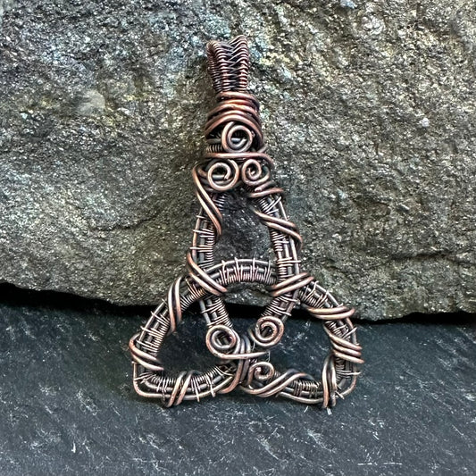 A copper-wrapped wire pendant shaped like a stylized triquetra or Celtic knot, displayed against a textured gray rock background.