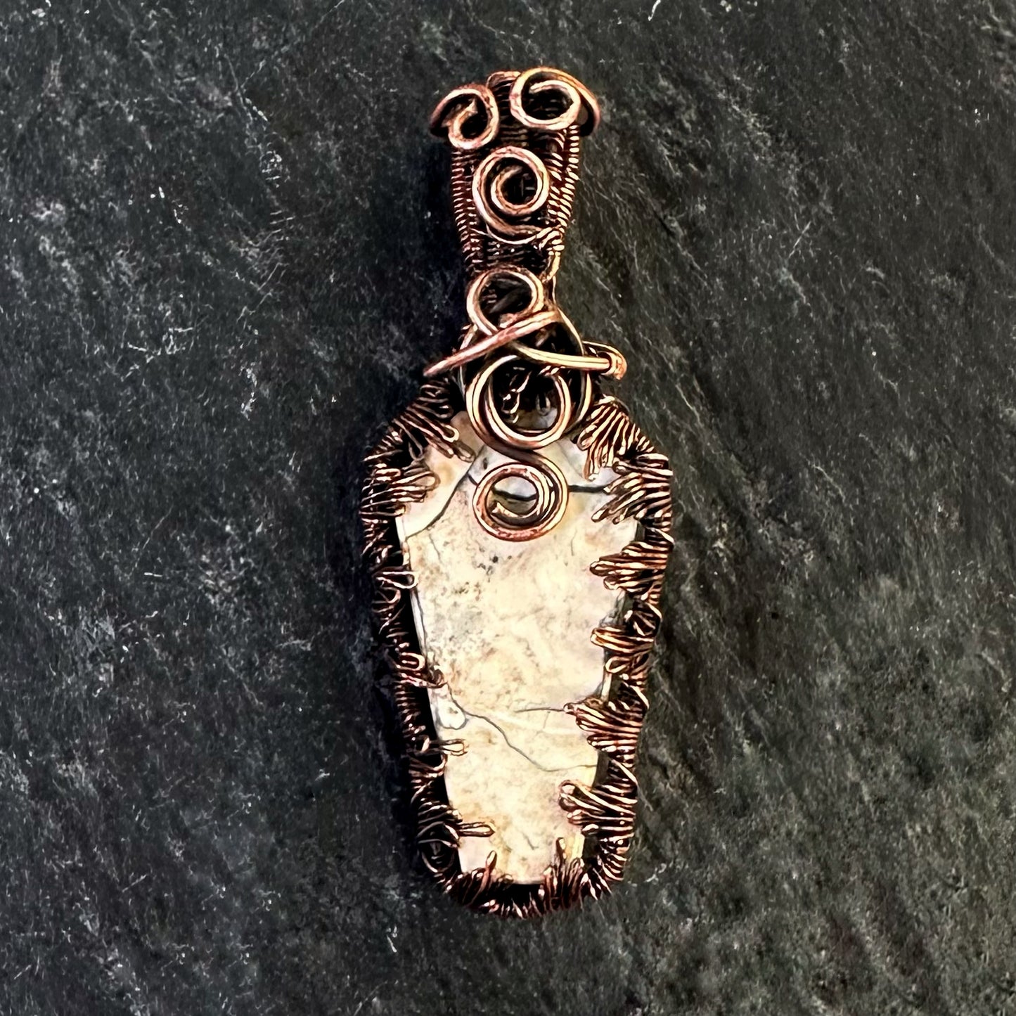 A handmade copper wire-wrapped coffin shaped pendant featuring a light coloured Maligano jasper stone, displayed on a dark textured slate background.