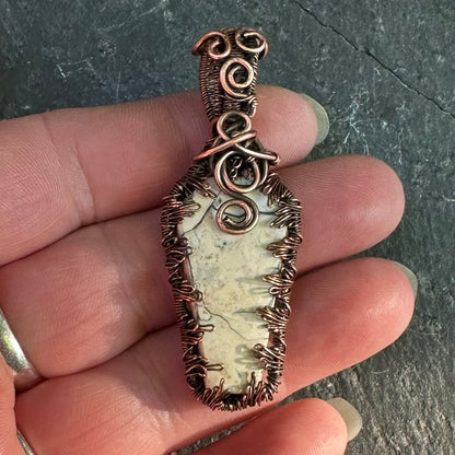 A handmade wire-wrapped pendant featuring a light coloured Maligano jasper with dark veining, held in a person's hand against a dark slate background.