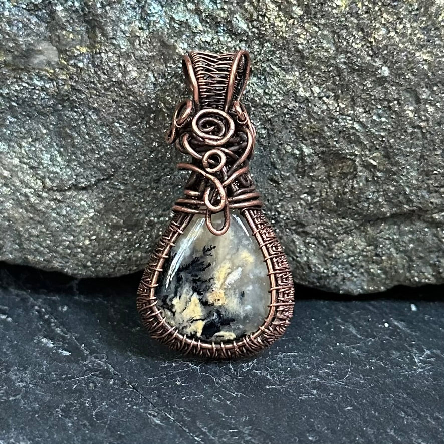 A teardrop-shaped pendant featuring a yellow and black dendritic agate stone wrapped in textured copper wire, set against a dark gray stone background.