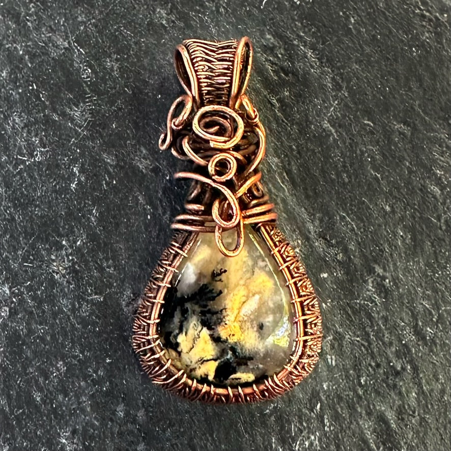 A handmade teardrop pendant featuring a marbled yellow, black, and white dendritic agate stone wrapped in detailed copper wire, displayed on a dark textured slate background.