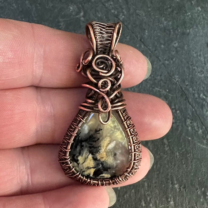 A close-up of a hand holding a copper-wrapped teardrop pendant with a yellow black dendritic agate stone, set against a dark slate background.