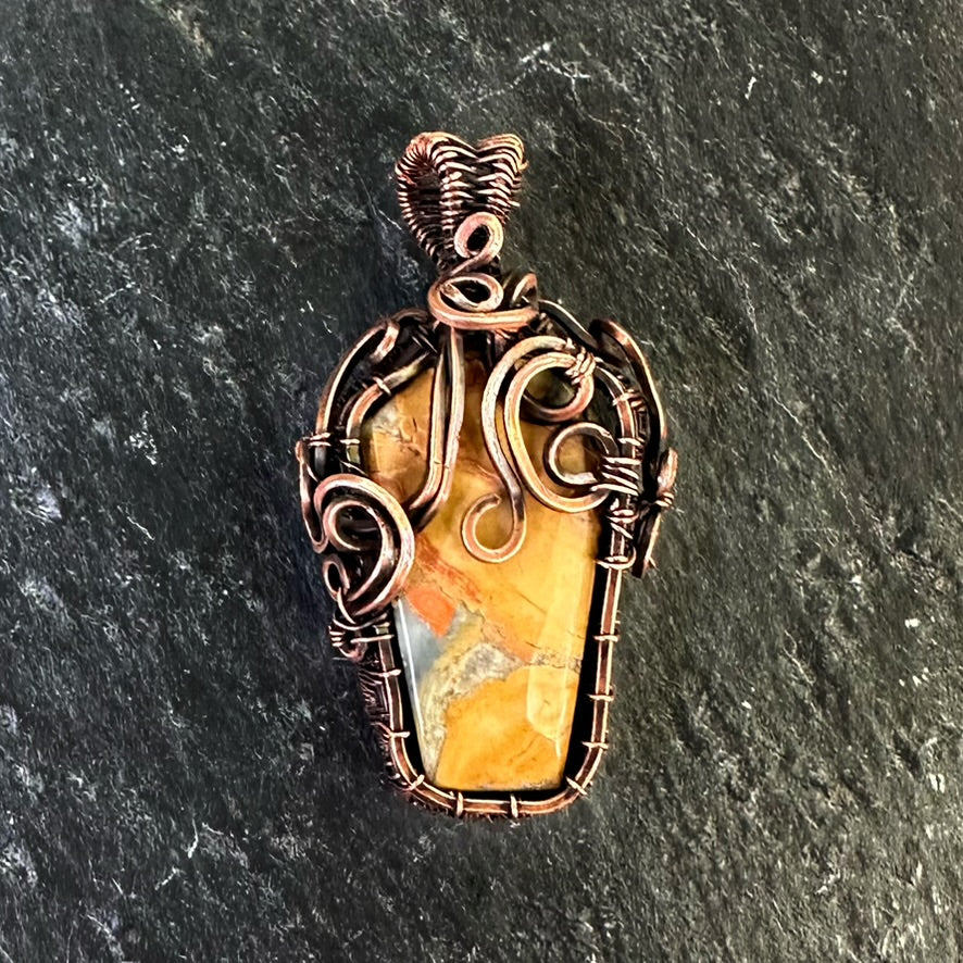 A handmade copper wire pendant featuring a polished coffin-shaped orange and yellow jasper stone, set against a dark textured slate background.