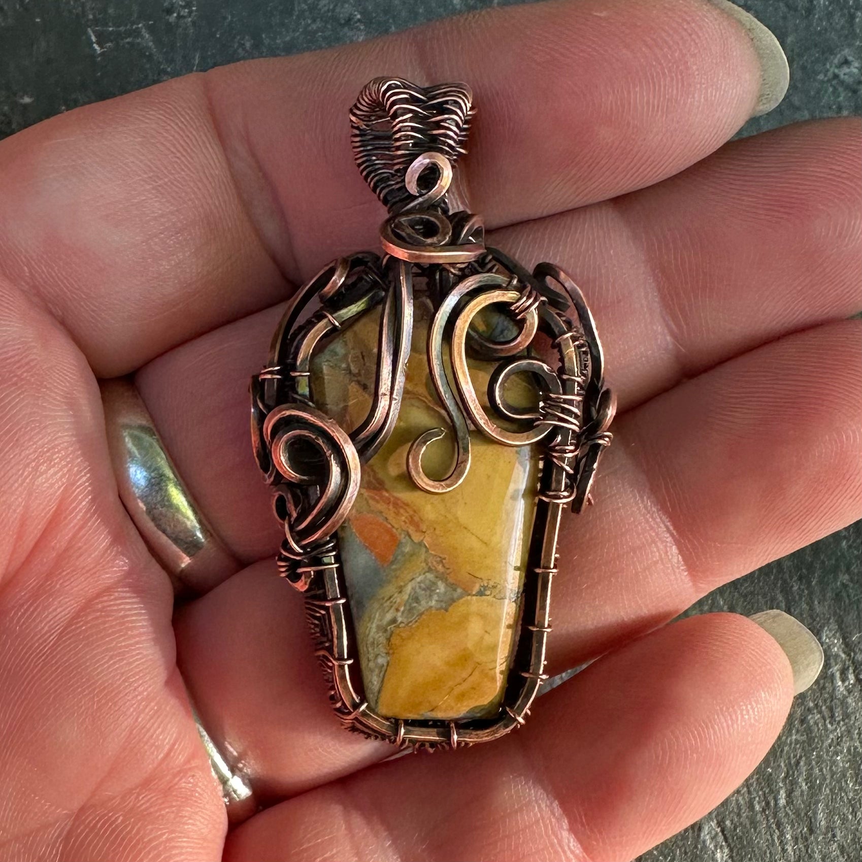 A close-up of a hand holding a gothic-style pendant featuring a coffin-shaped yellow and orange jasper stone wrapped in textured copper wire.