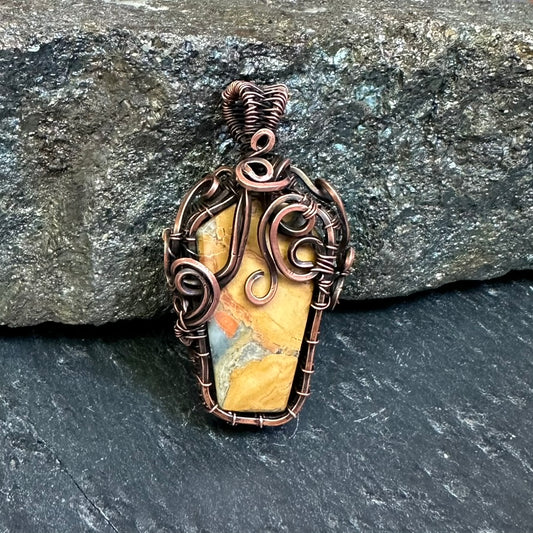 A copper wire-wrapped pendant featuring a polished coffin-shaped yellow and orange jasper stone, displayed against a dark textured slate and rock background.